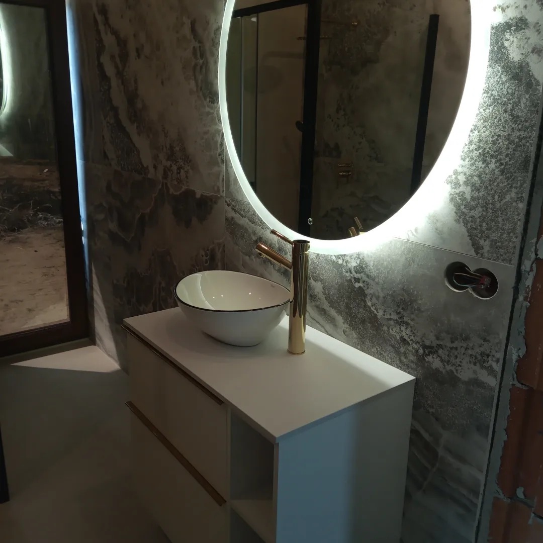 Nowoczesna łazienka z umywalką nablatową, złotą armaturą i okrągłym lustrem z podświetleniem LED; widoczne niedokończone wykończenie ściany z cegły.