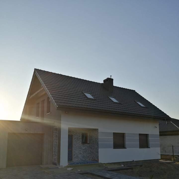 Nowo wybudowany dom jednorodzinny z szarą dachówką, trzema oknami dachowymi, częściowo wykończoną elewacją w odcieniach szarości i białego oraz garażem z bramą segmentową.