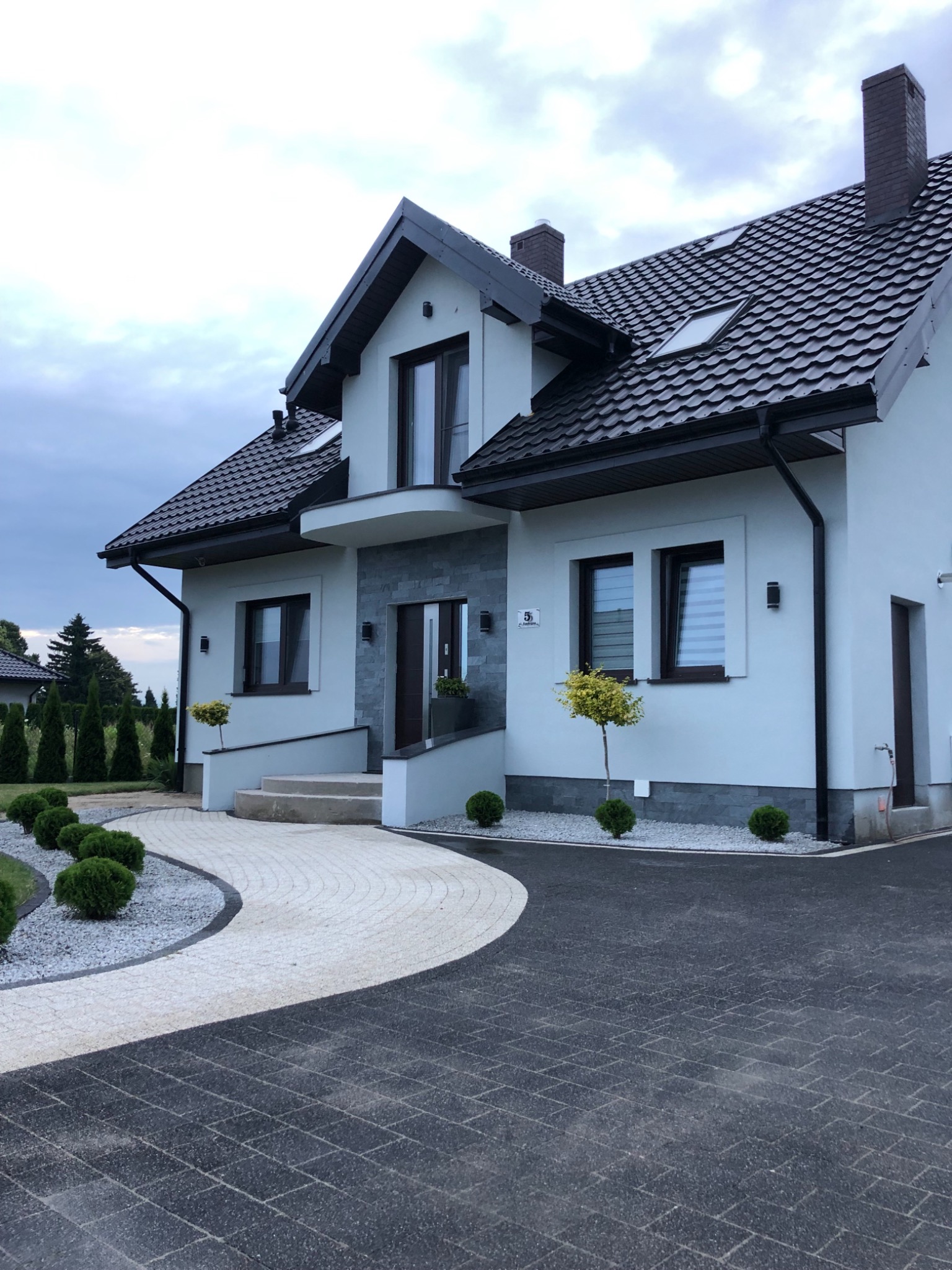 Elewacja jasnego domu jednorodzinnego z ciemną dachówką, ozdobnymi krzewami i dwukolorową ścieżką z kostki brukowej prowadzącą do wejścia.
