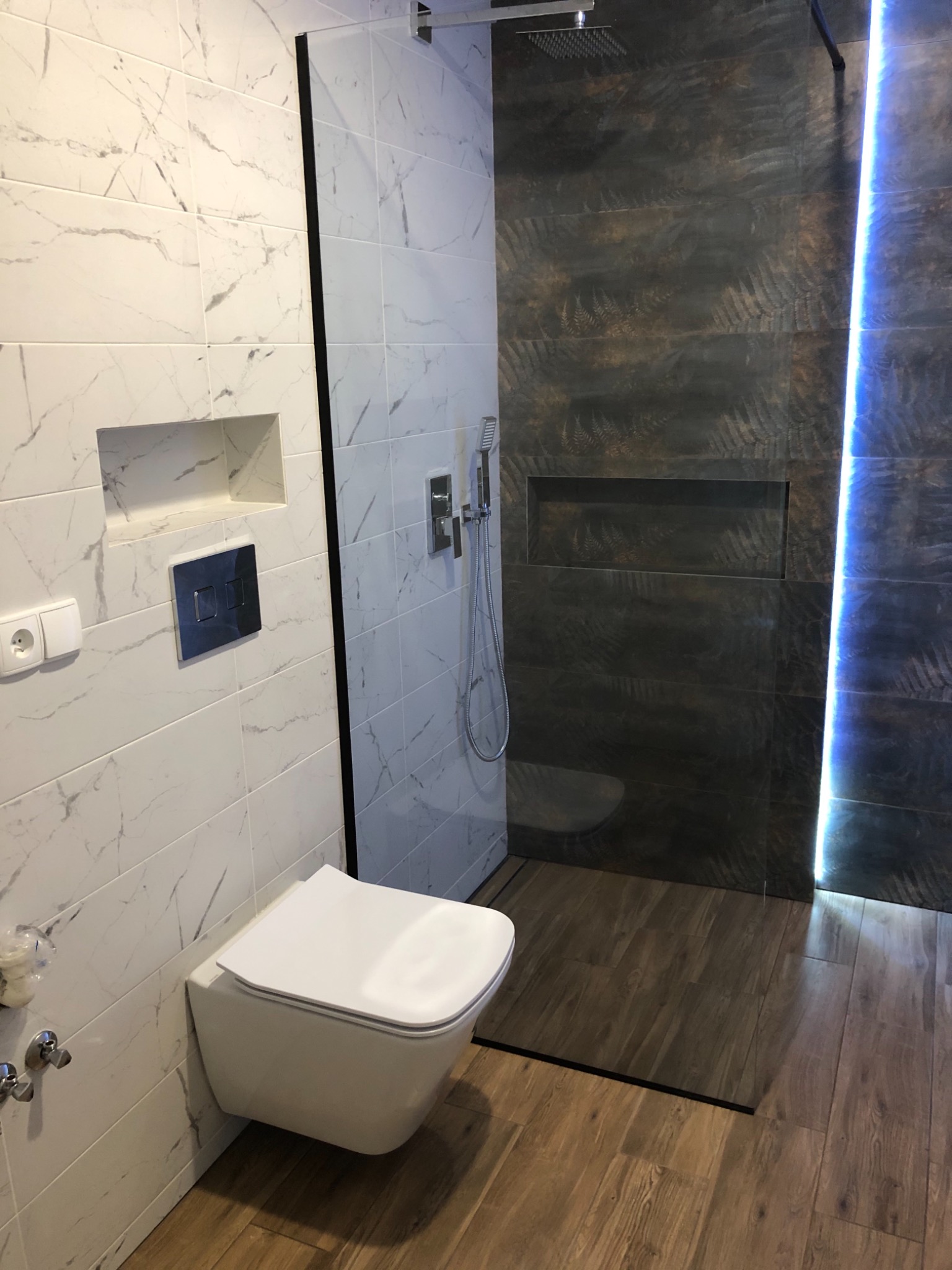 Nowoczesna łazienka z podwieszaną toaletą, marmurowymi płytkami, kabiną prysznicową typu walk-in z ciemnymi płytkami z motywem paproci i podświetleniem LED, oraz drewnopodobną podłogą.