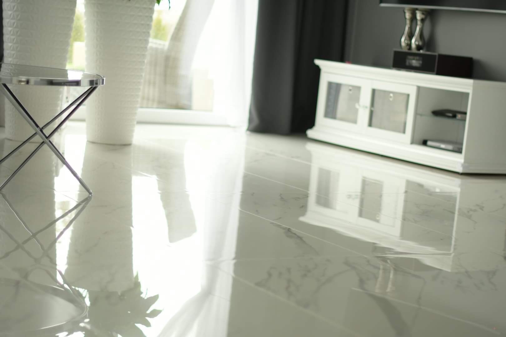 Błyszcząca, jasna podłoga z marmurowym wzorem, odbijająca światło i elementy wystroju: stolik pomocniczy, donice i biała szafka RTV.