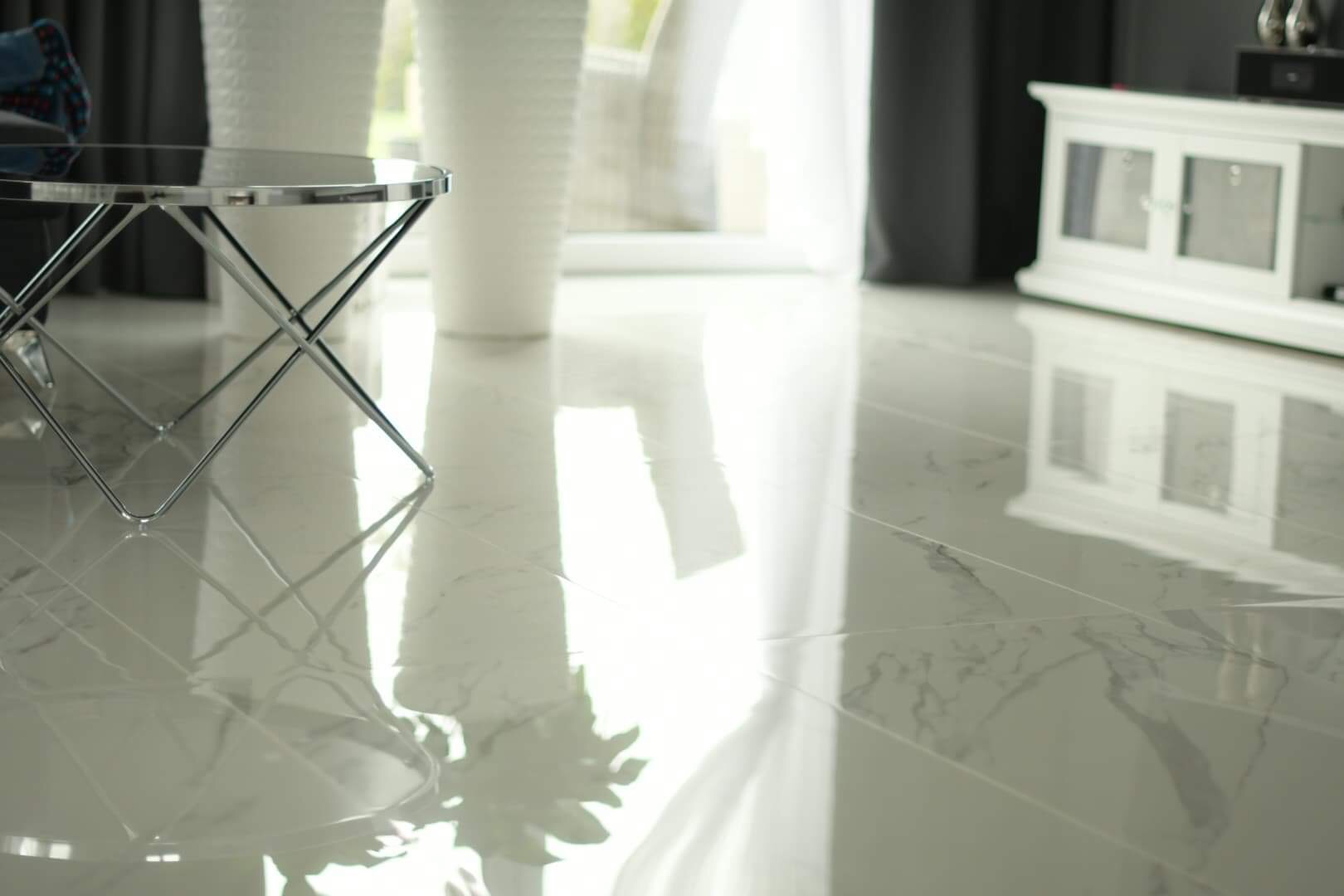 Błyszcząca, marmurowa podłoga w jasnym salonie z chromowanym stolikiem kawowym i białymi donicami w tle, odbijająca światło okna.