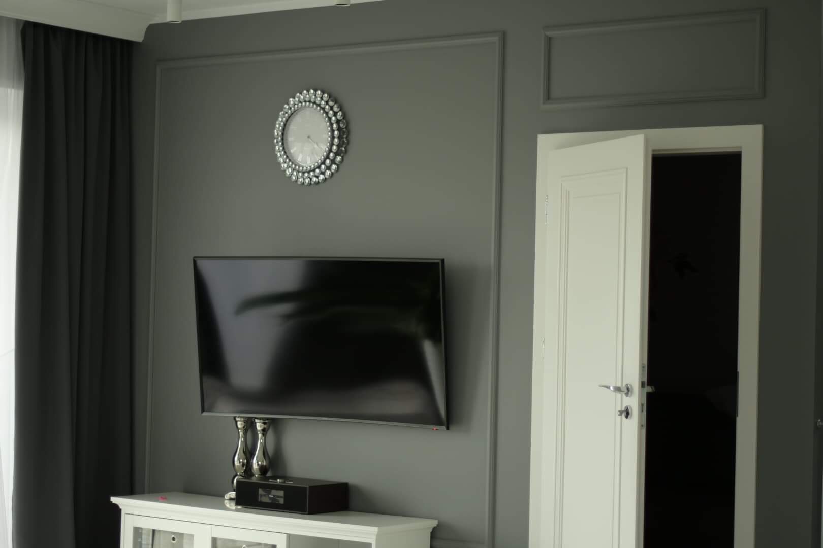 Szare wnętrze pokoju z telewizorem na ścianie z ozdobnymi listwami, zegarem z kryształkami i częściowo otwartymi białymi drzwiami.