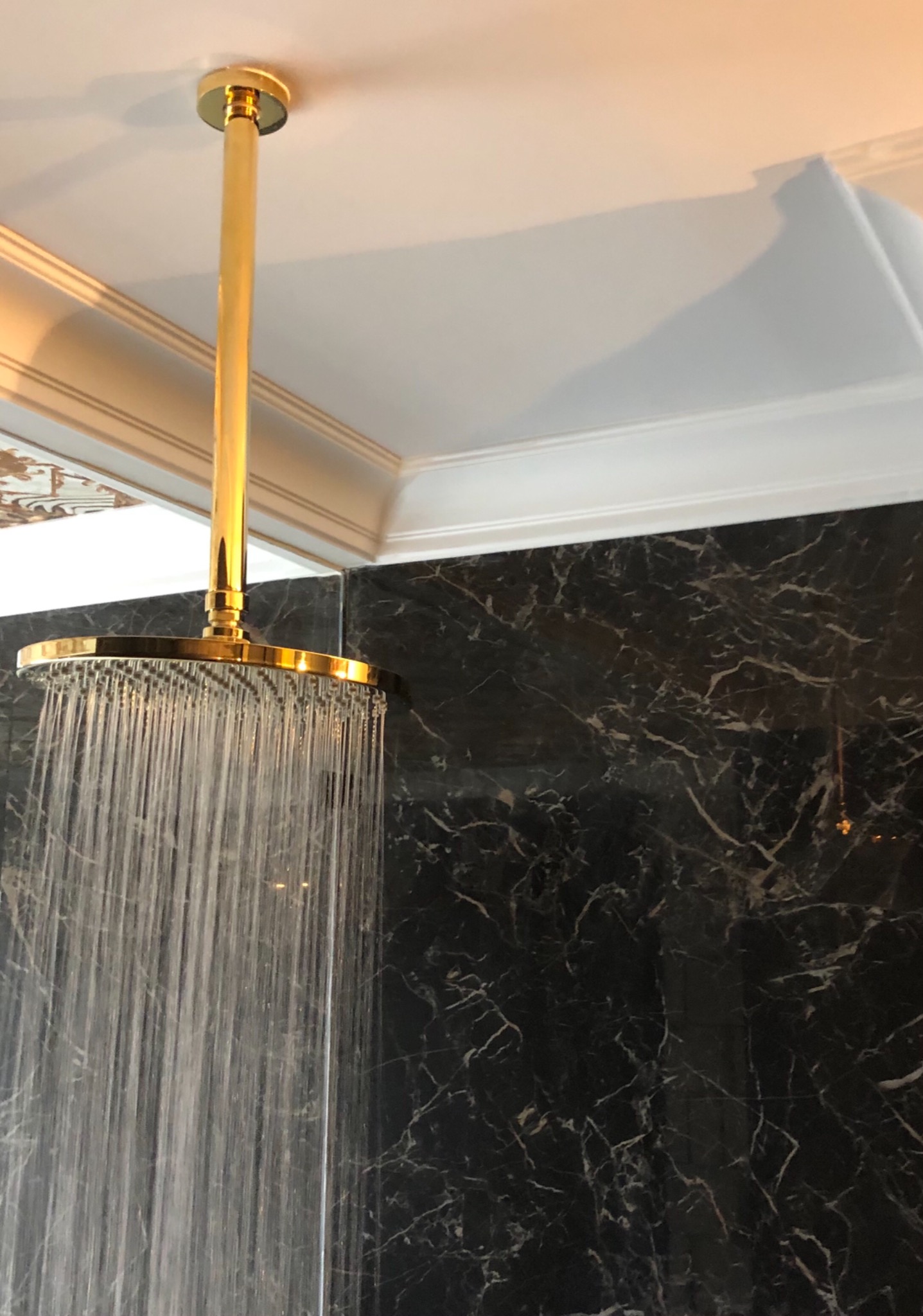 Deszczownica w kolorze złotym z włączonym strumieniem wody na tle ściany z czarnego marmuru z białymi żyłami, widoczny fragment sufitu z ozdobnym sztukaterią.
