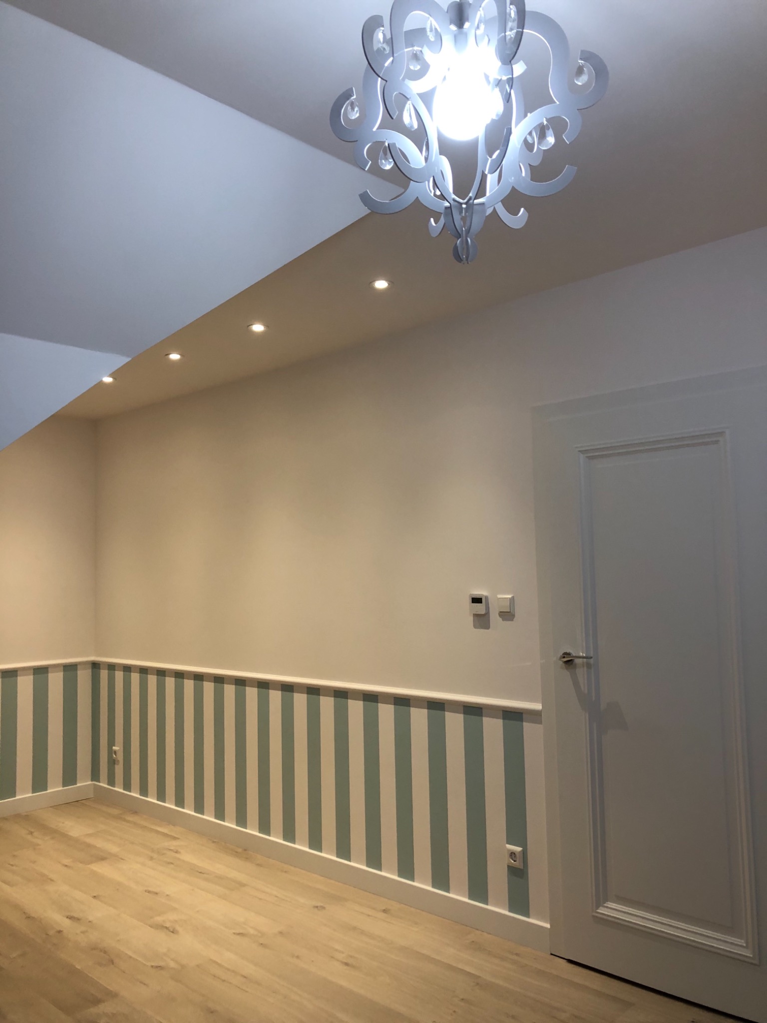 Pomalowane na biało ściany z dolnym pasem w pionowe, pastelowe paski, jasna podłoga z paneli, białe drzwi i ozdobny, biały żyrandol z żarówką, w pokoju po remoncie.