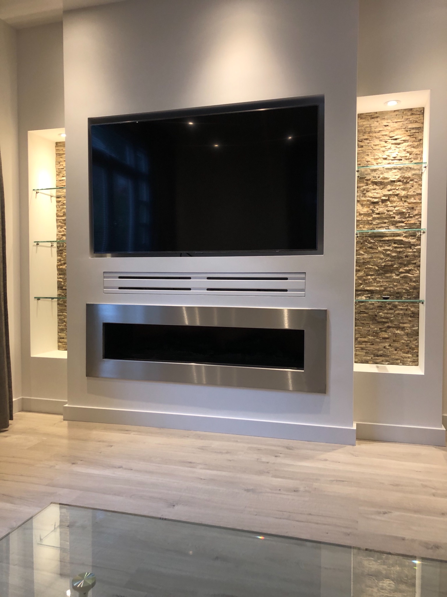 Nowoczesny salon z wbudowanym telewizorem, kominkiem elektrycznym w stalowej obudowie i kamiennymi wnękami z szklanymi półkami.