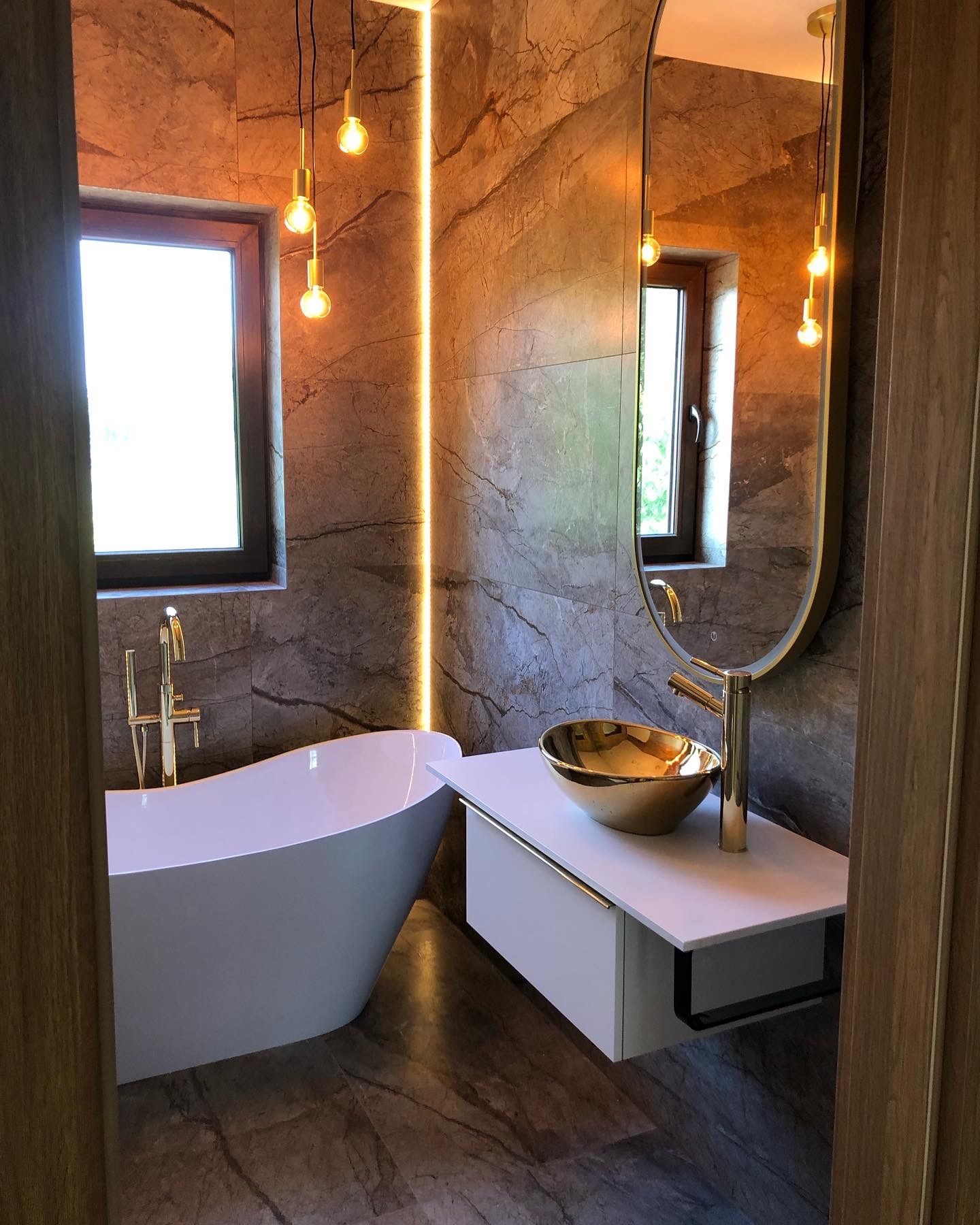 Elegancka łazienka z marmurowymi ścianami, wolnostojącą białą wanną, złotą armaturą i umywalką w kształcie misy, oświetlona wiszącymi żarówkami Edison i ledowym paskiem.