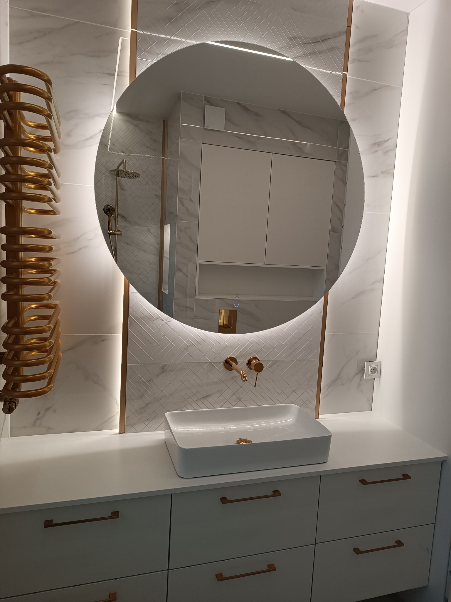 Elegancka łazienka z okrągłym lustrem podświetlanym LED, białą umywalką nablatową, złotą armaturą i grzejnikiem drabinkowym w kolorze złota na tle marmurowych płytek.