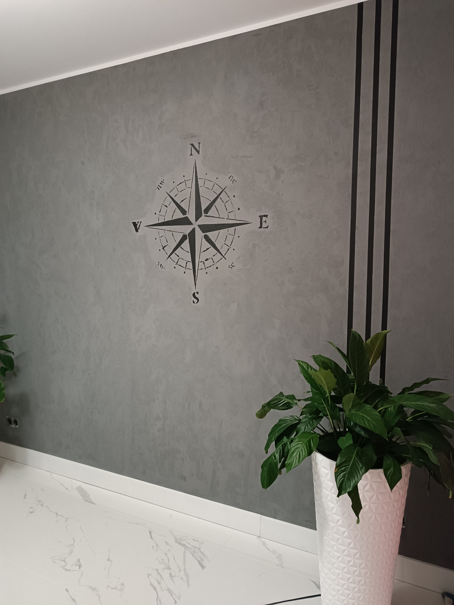 Szara ściana z dekoracyjną fakturą, ozdobiona motywem kompasu i trzema pionowymi czarnymi pasami, uzupełniona roślinami w białych donicach na marmurowej podłodze.