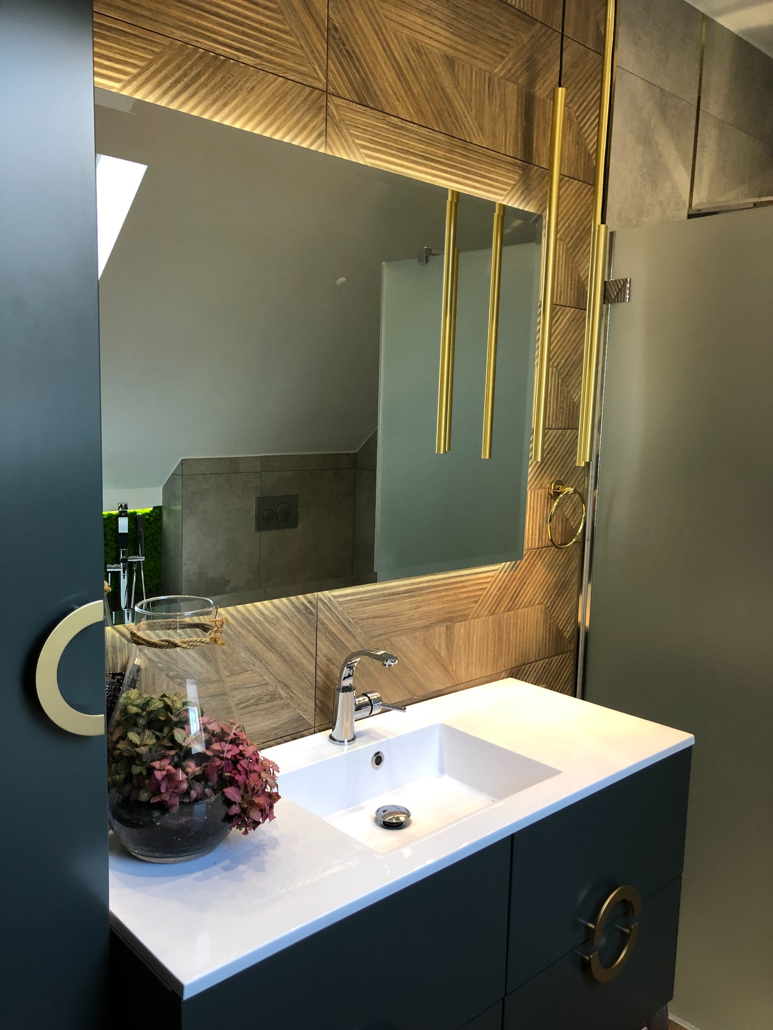 Nowoczesna łazienka w odcieniach szarości i drewna z umywalką nablatową, lustrem z podświetleniem LED i dekoracyjnymi złotymi elementami.