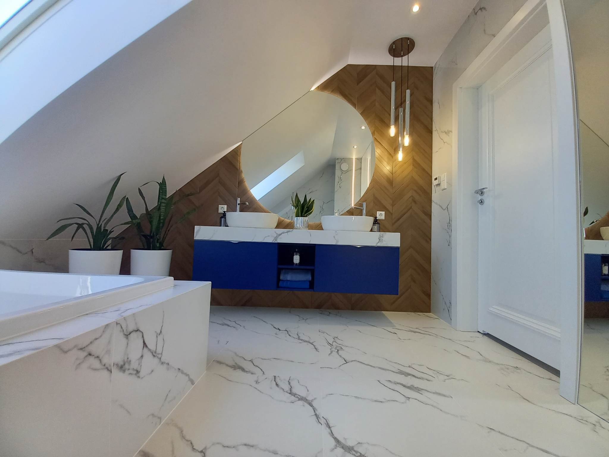 Nowoczesna łazienka na poddaszu z marmurową posadzką i okładziną w strefie umywalek, owalne lustro z podświetleniem, niebieska szafka pod umywalki i wiszące lampy.