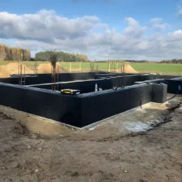 Zalane fundamenty budynku z hydroizolacją w postaci czarnej folii kubełkowej, wystające pręty zbrojeniowe, widok na plac budowy z hałdami ziemi i drzewami w tle, pochmurne niebo.