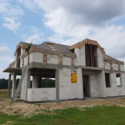 Surowy stan dwukondygnacyjnego domu z białych bloków, widoczne drewniane elementy konstrukcyjne dachu i stropu, na ścianie żółta tablica informacyjna, obok drabina, w tle pole i las.