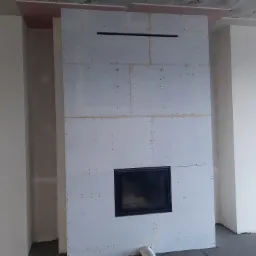 Konstrukcja ściany z płyt gipsowo-kartonowych z wbudowanym wkładem kominkowym, widoczne łączenia płyt i otwór wentylacyjny u góry, zarys słupów po bokach.