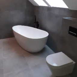 Nowoczesna łazienka z wolnostojącą białą wanną, podwieszaną toaletą i szarymi płytkami imitującymi beton, oświetlona przez okno dachowe.