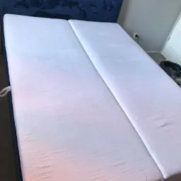 Podwójne łóżko z białym materacem i niebieskim zagłówkiem, widoczne gniazdko elektryczne w tle.