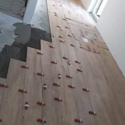 Luk-Ar Bud - Układanie podłogi z płytek imitujących drewno, z użyciem krzyżyków dystansowych, widoczny fragment świeżo nałożonego kleju i przygotowane paczki płytek.