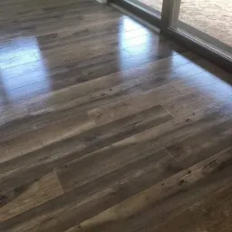 Ułożone panele podłogowe o wzorze imitującym drewno, widoczne odbicia światła, przy oknie tarasowym z widokiem na zewnątrz.