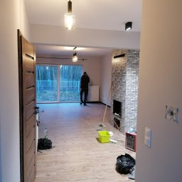 Luk-Ar Bud - Wnętrze po remoncie z podłogą imitującą drewno, kominkiem z szarej cegły i widokiem na las przez duże okno; widoczny pracownik w oddali.