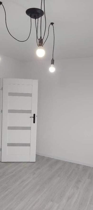 Wnętrze pokoju po remoncie: białe ściany, jasne panele podłogowe, białe drzwi z poziomymi przeszkleniami i czarna lampa wisząca z kilkoma żarówkami na długich kablach.