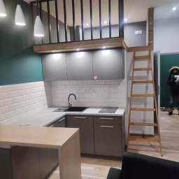 Łukasz - Nowoczesna kuchnia z antresolą i drewnianą drabinką, szaro-białe szafki, białe podłużne płytki na ścianie, ciemnozielona ściana w tle, minimalistyczne lampy wiszące, widoczny fragment sofy