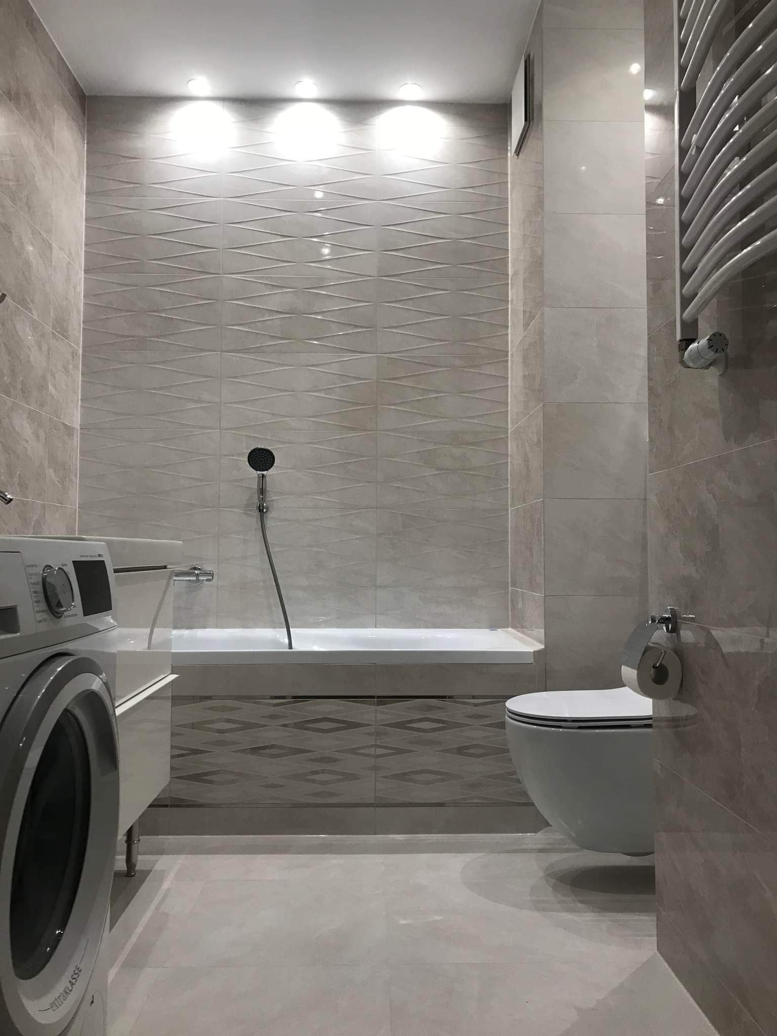 Jasna łazienka z pralką, toaletą i wanną obudowaną płytkami z geometrycznym wzorem; ściany wyłożone płytkami w odcieniach beżu i szarości, oświetlenie punktowe sufitowe.