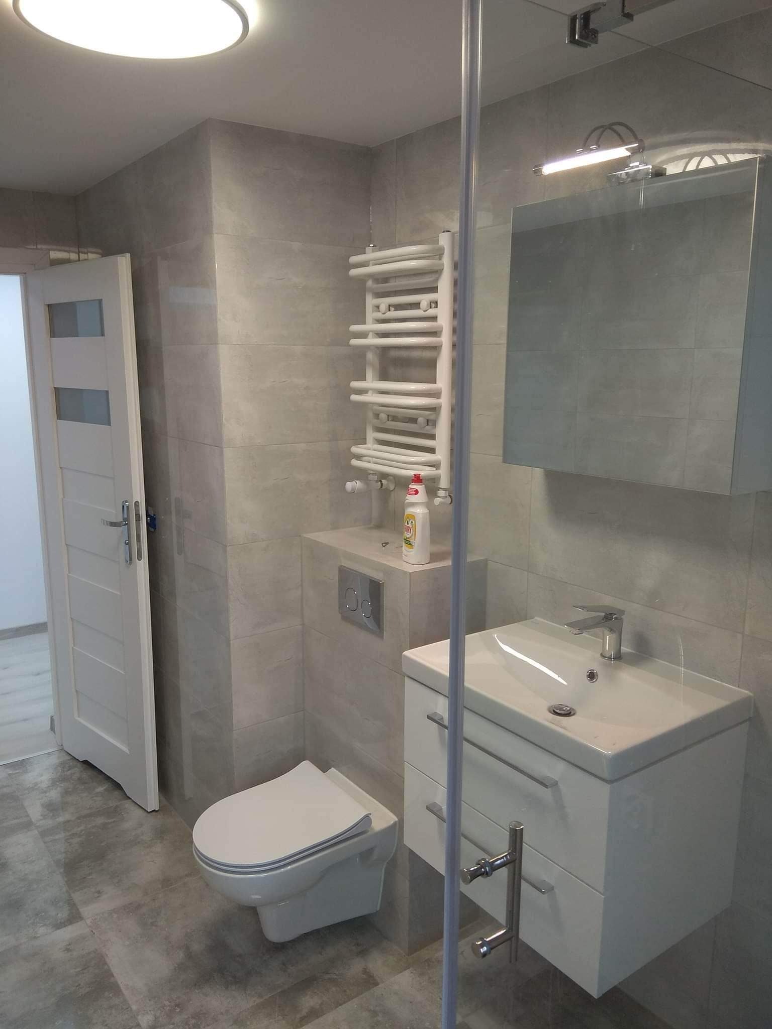 Nowoczesna łazienka w szarościach z podwieszaną toaletą, grzejnikiem drabinkowym i szafką z umywalką, widoczna butelka płynu do mycia naczyń.