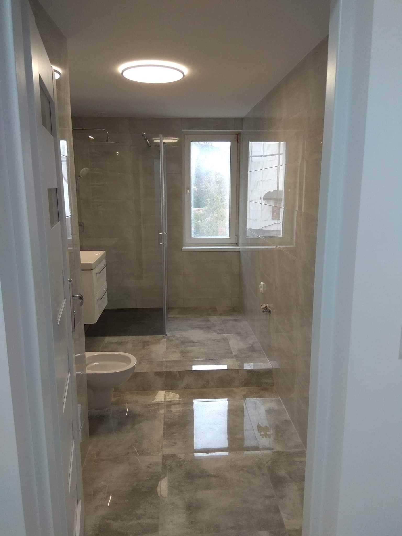 Nowoczesna łazienka z szarymi płytkami, kabiną prysznicową typu walk-in, podwieszaną toaletą i oknem z widokiem na zieleń.