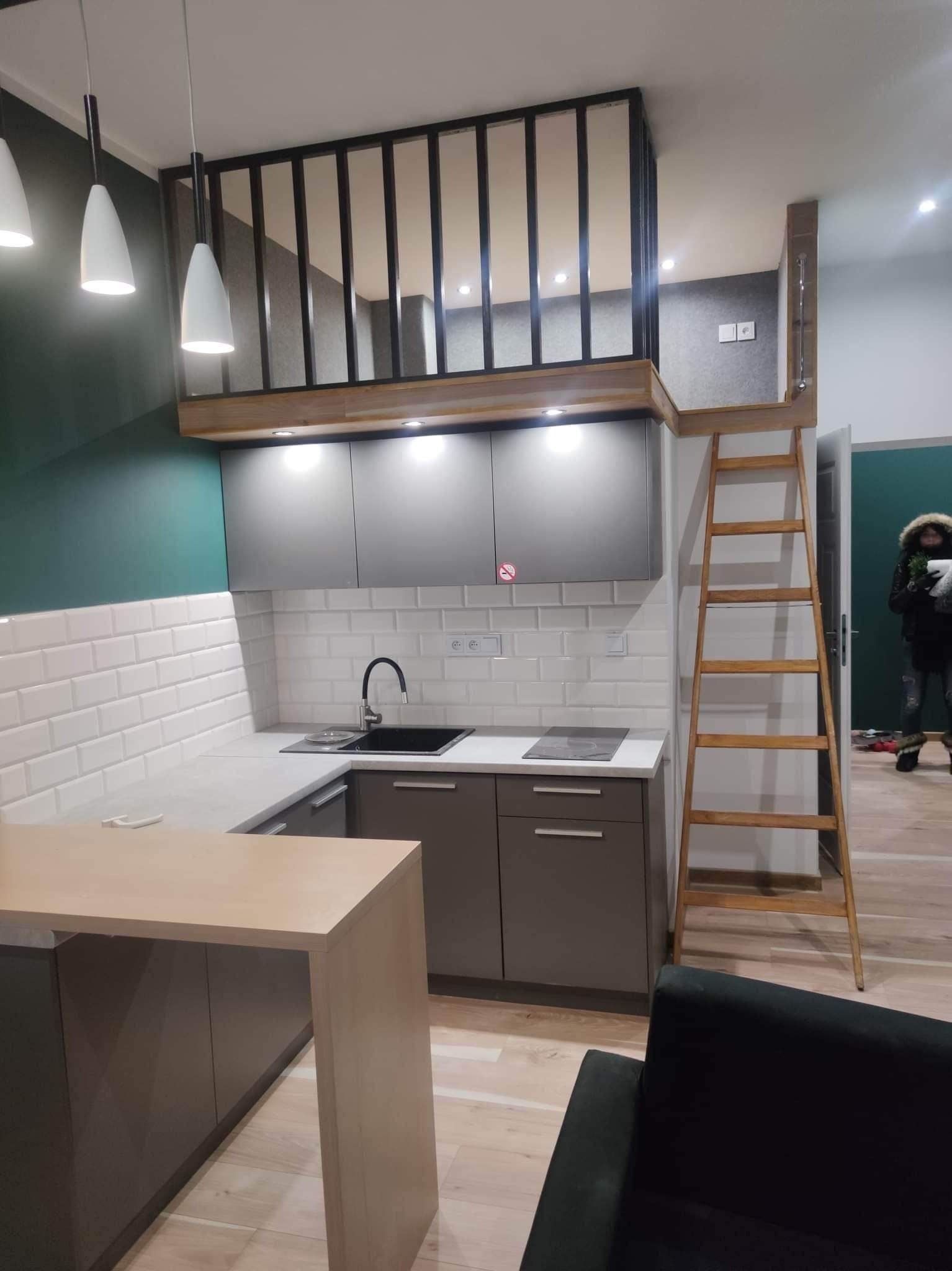 Nowoczesna kuchnia z antresolą i drewnianą drabinką, szaro-białe szafki, białe podłużne płytki na ścianie, ciemnozielona ściana w tle, minimalistyczne lampy wiszące, widoczny fragment sofy