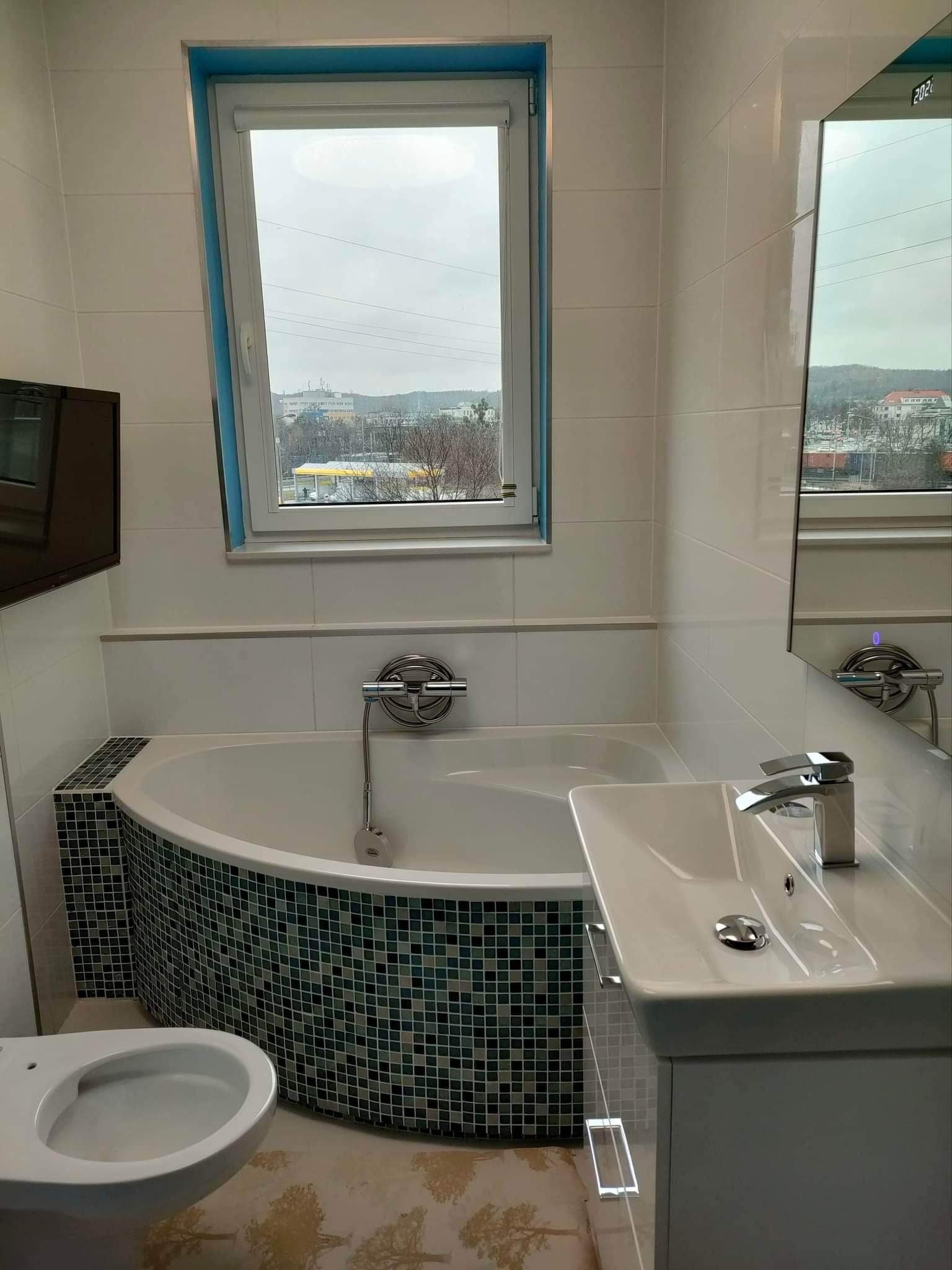 Wykończona łazienka z białą armaturą, mozaiką na obudowie wanny i widokiem na miasto za oknem.