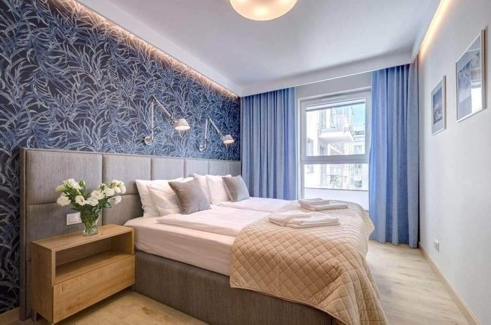 Nowoczesna sypialnia z tapetą w motywy roślinne, oświetleniem punktowym nad łóżkiem, jasnymi zasłonami i drewnianą szafką nocną z bukietem białych kwiatów.