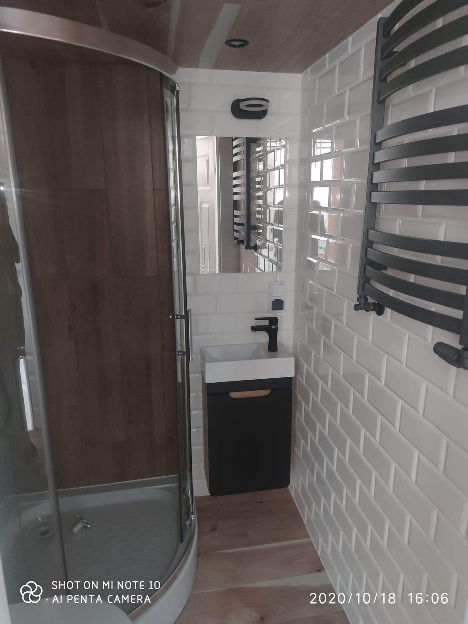 Nowoczesna łazienka z kabiną prysznicową z brązowymi panelami, białą umywalką nablatową z czarną armaturą, grzejnikiem drabinkowym i białymi, błyszczącymi płytkami typu metro