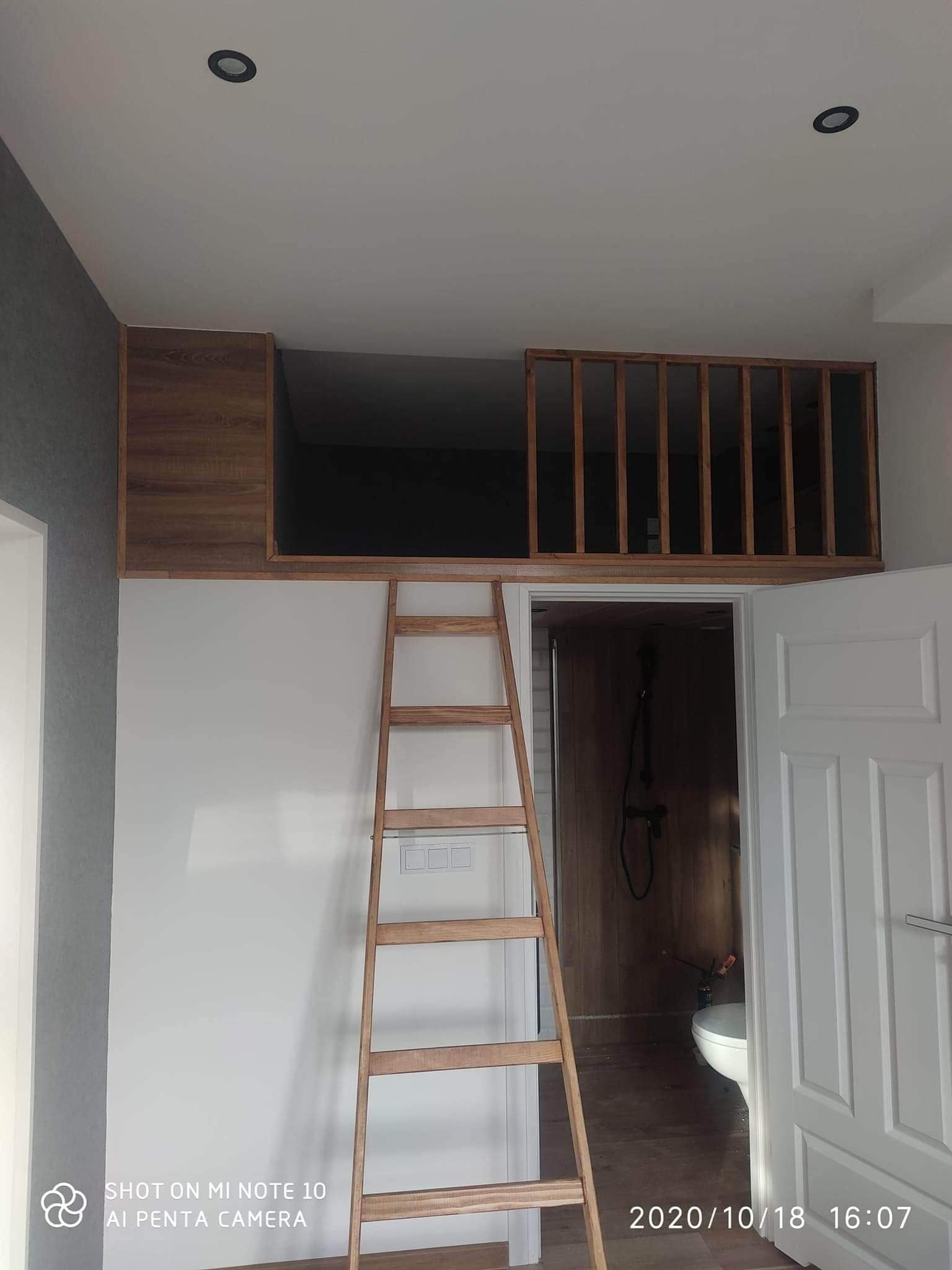 Wysoka łazienka z antresolą, drewnianą drabinką i zabudową nad drzwiami, widok na kabinę prysznicową z drewnianymi panelami.