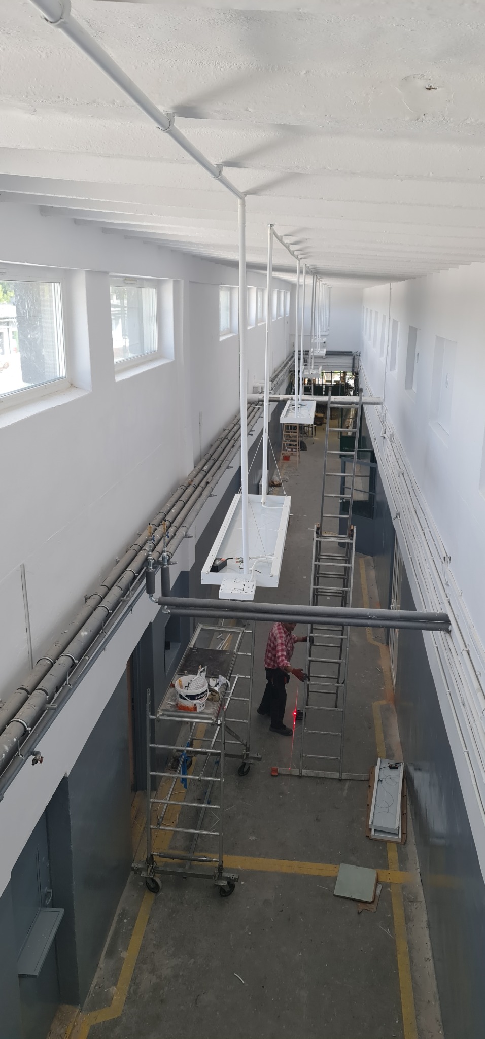 Wysoki kadr z góry przedstawia montaż lamp w długim, białym korytarzu z rurami pod sufitem; pracownik na rusztowaniu ustawia laserowy poziom.