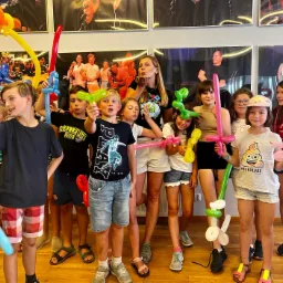 Grupa dzieci z balonami w kształcie zwierząt i mieczy, prowadzona przez animatorkę, pozuje do zdjęcia na tle ściany z czarno-białymi portretami.