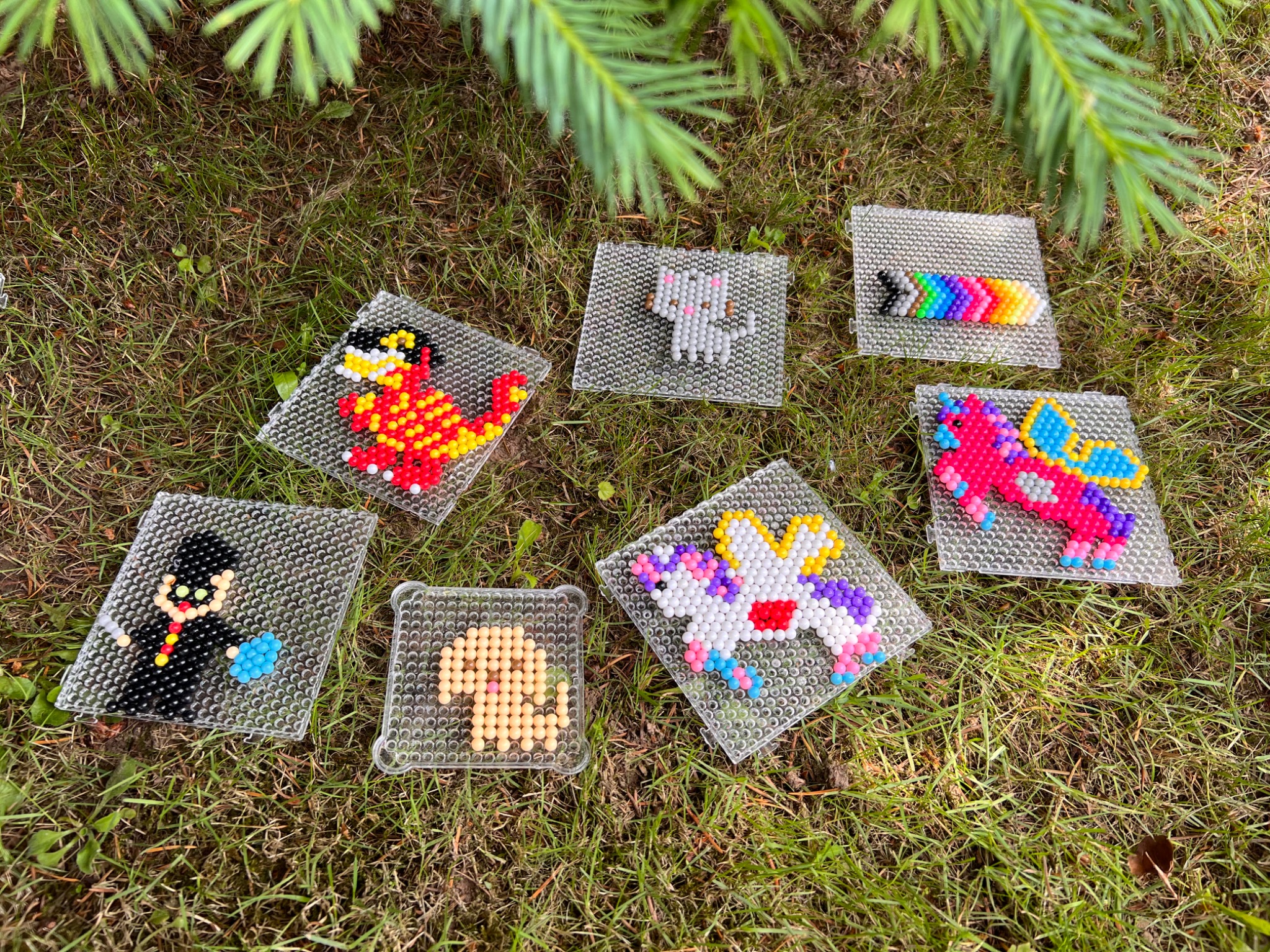 Ręcznie robione, kolorowe breloczki z koralików Hama ułożone na trawie: smok, słoń, tęcza, Harry Potter, pies i dwa jednorożce z pegazimi skrzydłami, na przezroczystych podkładkach.