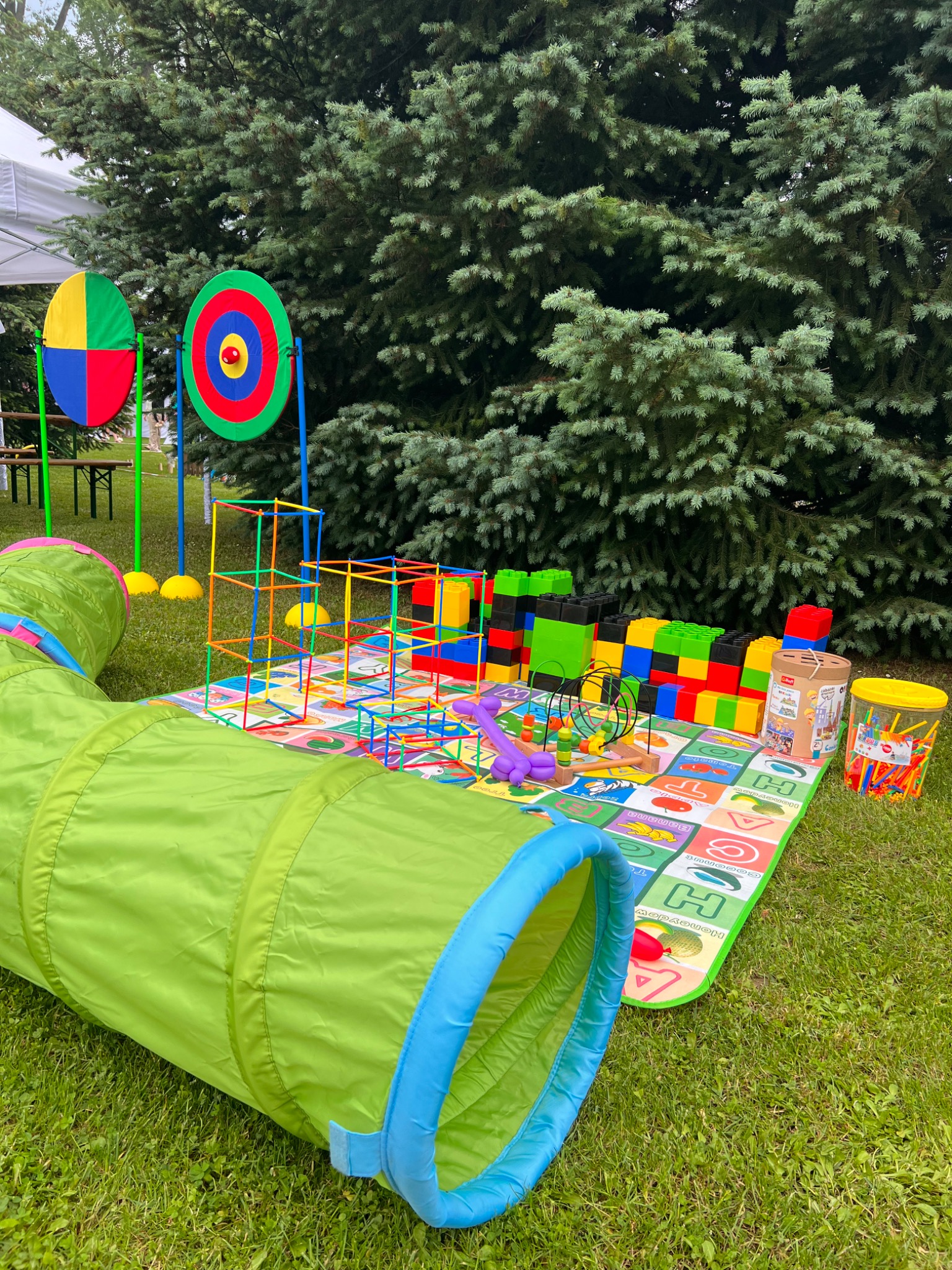 Kolorowy kącik zabaw dla dzieci na trawie w Warszawie, z tunelem, dużymi klockami, konstrukcjami z rurek i matą edukacyjną z literami, na tle zielonych drzew.