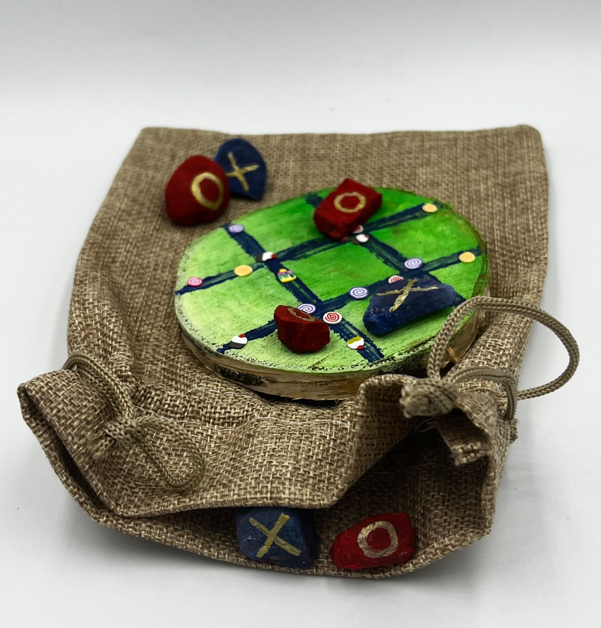 Ręcznie wykonana gra w kółko i krzyżyk z drewnianą planszą pomalowaną na zielono z niebieskimi liniami i kolorowymi detalami, kamienie do gry w kształcie jajek leżące na jutowym woreczku.