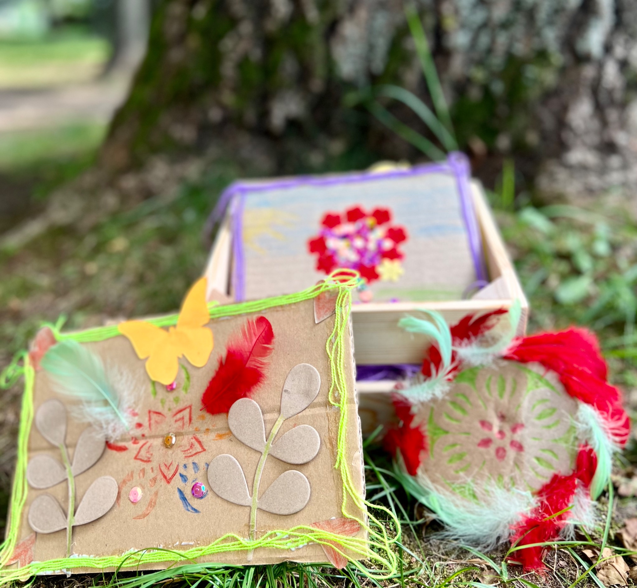 Ręcznie robione ozdoby z kartonu, piór i cekinów, leżące na trawie przed pniem drzewa, w tym kartonowe pudełko z dekoracją w środku oraz kartka z naklejonym motywem roślinnym i piórami.