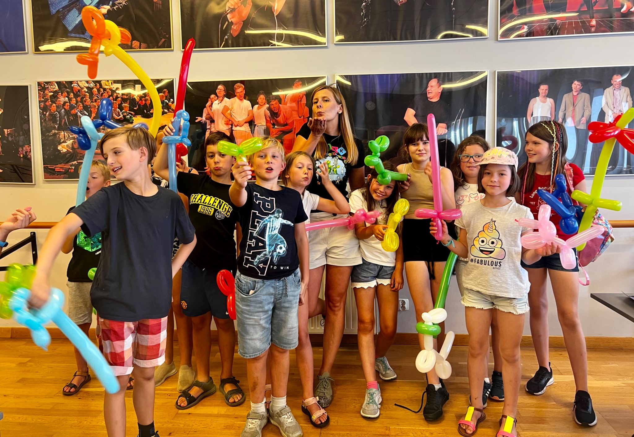Grupa dzieci z balonami w kształcie zwierząt i mieczy, prowadzona przez animatorkę, pozuje do zdjęcia na tle ściany z czarno-białymi portretami.