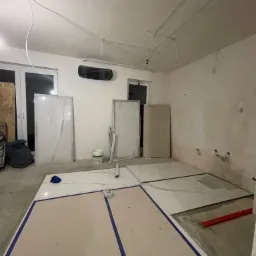 Wnętrze w trakcie kładzenia płytek podłogowych, z widocznymi płytami o wzorze marmuru, częściowo zakrytymi folią i taśmą malarską, oraz odsłoniętym fragmentem podłoża z poziomnicą.