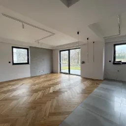 Wnętrze w trakcie wykończenia: parkiet ułożony w jodełkę kontrastuje z szarą, położoną częściowo podłogą z płytek. Widoczne okna z czarnymi ramami, sufit z oświetleniem szynowym i lampy wiszące.