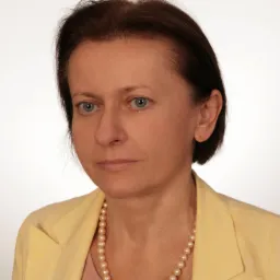 Portret kobiety o krótkich, brązowych włosach i zielonych oczach, ubrana w żółtą marynarkę i perłowy naszyjnik na jasnym tle.