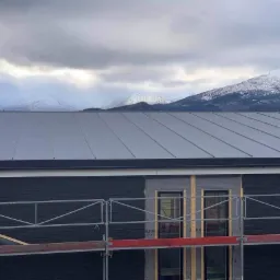 Nowy, szary dach panelowy na tle górskiego krajobrazu z ośnieżonymi szczytami, widoczne rusztowania.