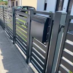 Nowoczesna, antracytowa brama wjazdowa z poziomymi panelami, zintegrowana ze skrzynką na listy, na tle jasnego domu jednorodzinnego i brukowanego chodnika.