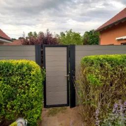 Nowoczesna furtka z paneli kompozytowych w kolorze szaro-beżowym, z czarną ramą, wkomponowana w ogrodzenie panelowe, otoczona zielenią krzewów i kwiatów.