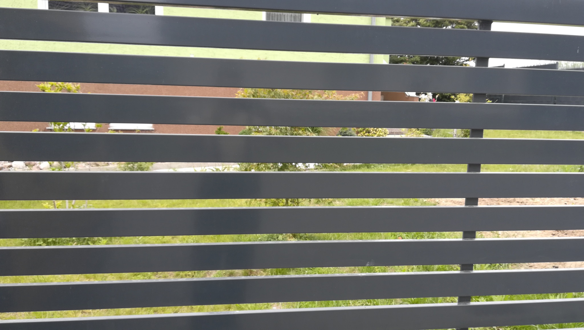 Szare poziome przęsła ogrodzeniowe z widokiem na trawnik i budynek w tle, ujęcie z bliska.