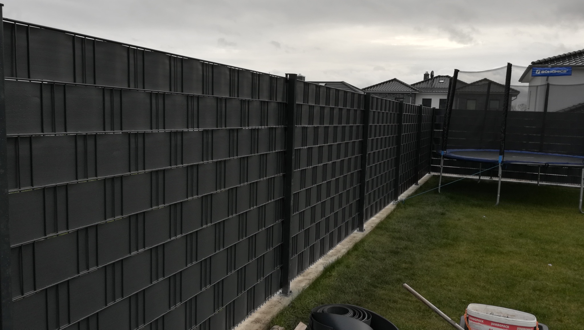 Nowoczesne ogrodzenie panelowe w kolorze antracytowym z widoczną konstrukcją i wypełnieniem, świeżo zamontowane na podmurówce, z elementami montażowymi i narzędziami w trawie w tle dom z trampoliną.