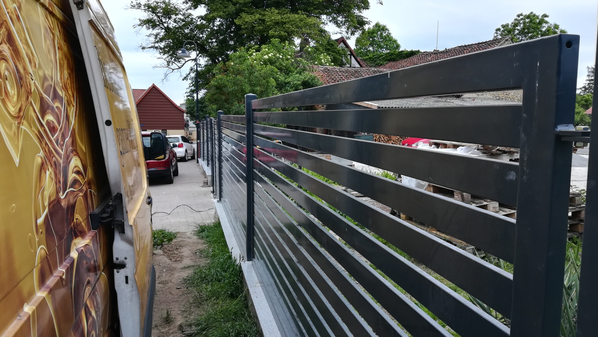 Nowoczesne, poziome przęsła ogrodzeniowe w kolorze grafitowym, widoczne z perspektywy ulicy w Gorzowie Wielkopolskim, obok zaparkowany samochód dostawczy z otwartymi drzwiami.