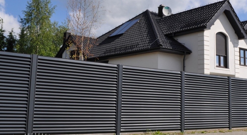 Nowoczesne ogrodzenie z poziomych, metalowych profili w kolorze grafitowym, na tle domu z ciemną dachówką i oknami z roletami zewnętrznymi.